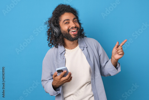 Young laughing cheerful Arabian man student in wireless headphones listens to audio podcast or watches stand-up on mobile phone and points finger towards stands in blue studio with smile