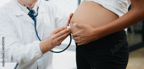 Pregnant african woman has appointment with doctor at clinic. Male gynaecologist OB GYN medic specialist with stethoscope listens to baby's heartbeat in mother's belly.