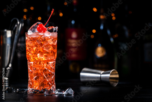 Rose with cherry, red cocktail drink with pink vermouth, amaretto liqueur, cherry juice, cocktail cherry and ice, black bar counter background, tools and bottles photo