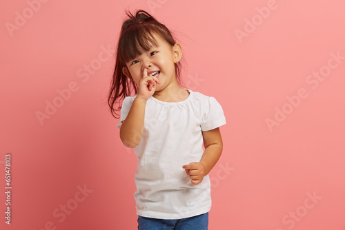 Portrait of little joyful Asian girl picking her nose on pink isolated background. Capricious and stubborn child does not obey his parents. Concept of child crisis of three years. photo