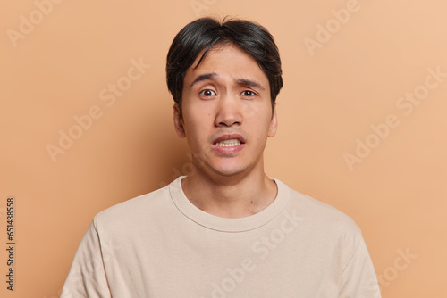 Human facial expressions concept. Brunet young Japanese man clenches teeth and looks embarrassed at camera dressed in casual t shirt dressed in casual t shirt isolated over brown background. © Wayhome Studio