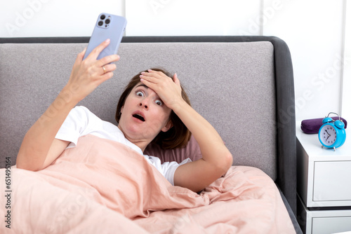 Young shocked woman waking up too late. Sleepy girl lying on the bed and holding mobile phone which ringing at the morning time