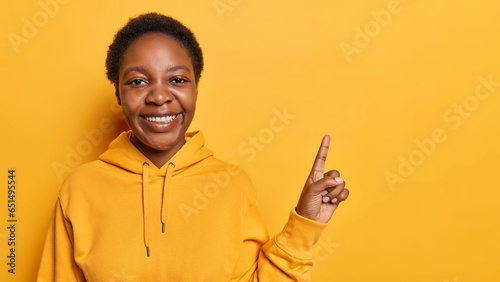 Joyful curly haired woman dressed in casual sweatshirt points index finger on empty space demonstrates advertisement dressed in casual hoodie isolated over yellow background promots discount