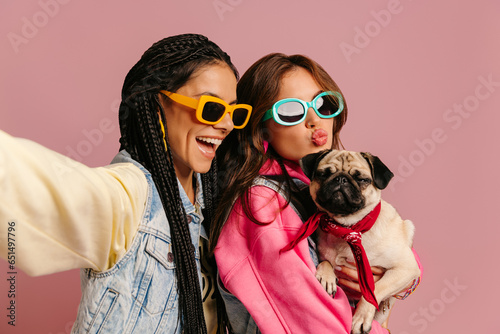 Two joyful young women in trendy glasses carrying cute pug dog and making selfie against pink background © gstockstudio