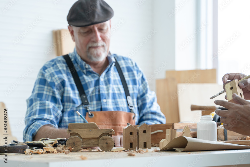 Selective focus on car and house wooden toy on table. Caucasian senior old white bearded man carpenter in apron and hat working in workshop, sketching on paper in white room Selective focus on car and house wooden toy on table. Caucasian senior old white bearded man carpenter in apron and hat working in workshop, sketching on paper in white room