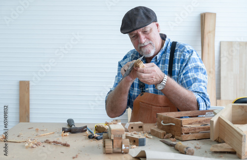 Caucasian senior old white bearded man carpenter in apron and hat working in workshop, use sandpaper polishing handmade wood toy, tools machine and small wooden model or handcrafts is on table