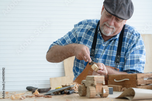 Caucasian senior old white bearded man carpenter in apron and hat using chisel working in workshop, tools machine small wooden toy model or handcrafts is on table in white room. Professional carpentry