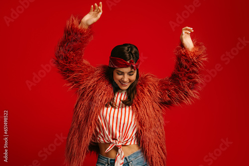 Stylish young hipster woman wearing fluffy coat and dancing on red background