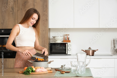 Young woman cooking chicken soup with cut carrot in kitchen
