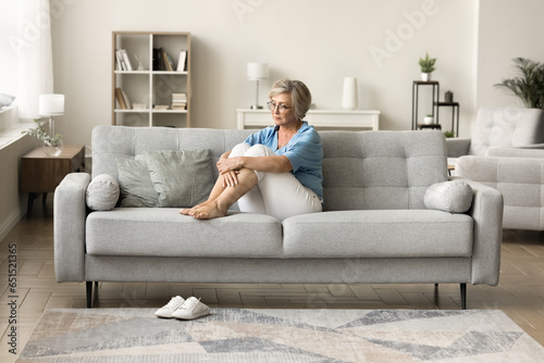 Depressed older retired woman sitting on sofa at cozy modern home, looking away in deep thoughts, staring into vacancy, thinking on depression, apathy, loneliness, remembering photo