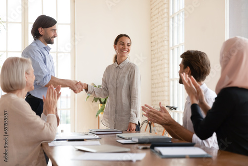Concept of the best employee promotion, praising or monetary bonus. Boss shake hands with successful team member mixed-race proud happy woman during meeting at boardroom while diverse staff applauding