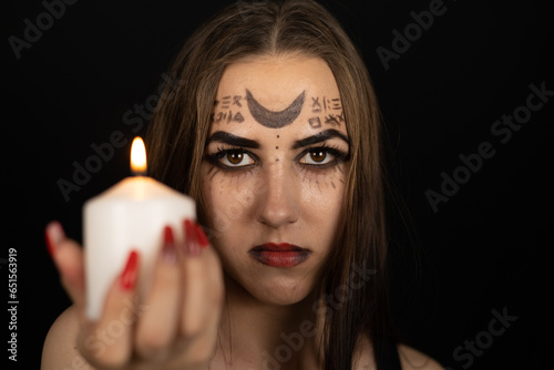 Looking at the camera, a witch with magical symbols on her face holds a burning candle. Halloween. In the dark, a beautiful witch looks askance at the camera. Halloween holiday concept. photo