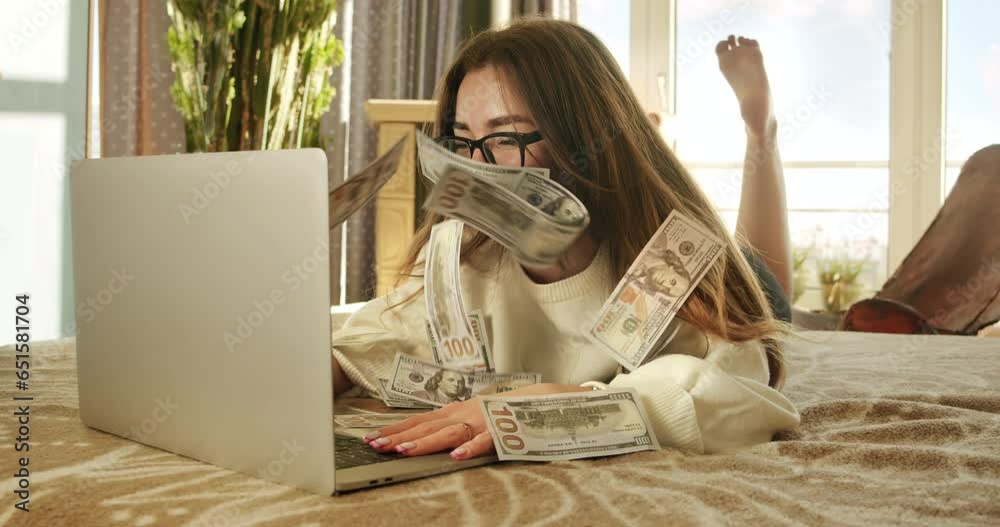 Cute girl making money online lying on couch, earn money. Cash flow. Online winning. Money rain flow through the laptop. Dollar bills floating in the student girl face