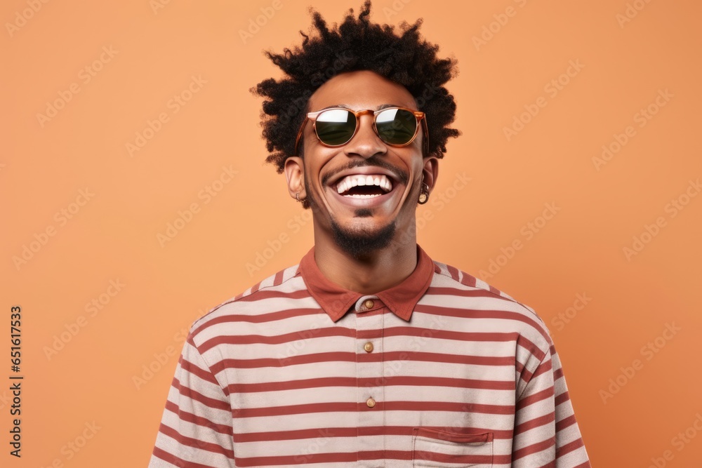 A suave man in sunglasses and a striped shirt stands against a wall, his jawline sharp and smile cool, exuding effortless style and confidence