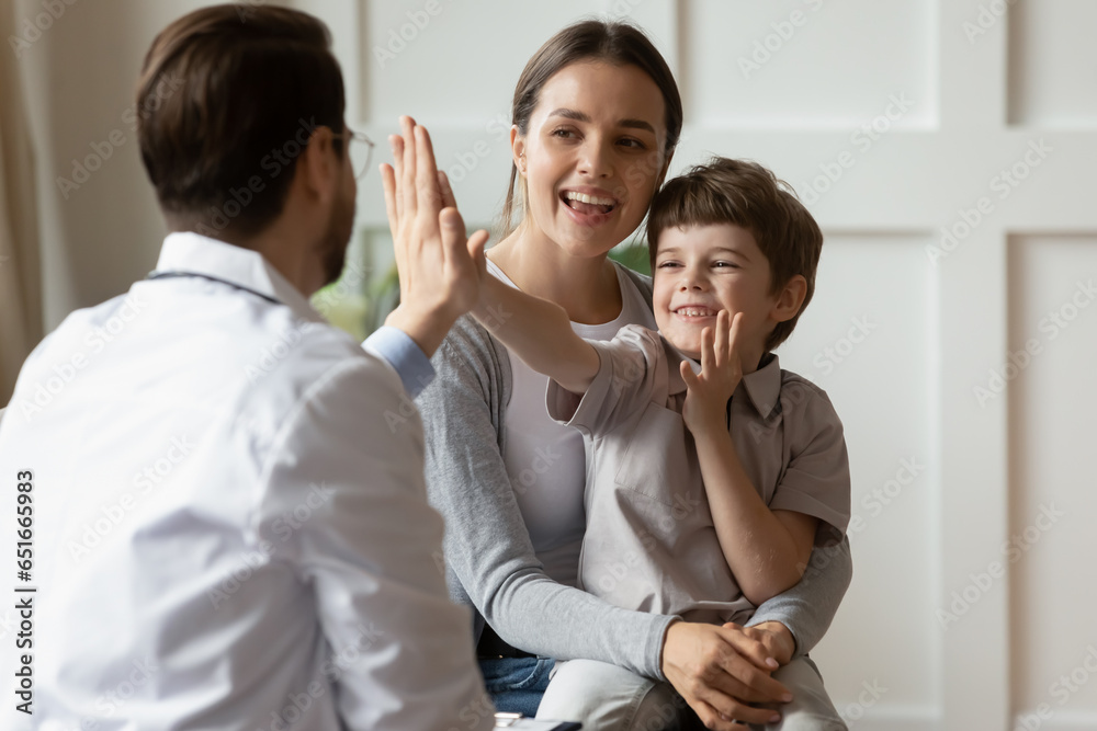 Happy cute little boy giving high five to pediatrician, friendly doctor greeting preschool patient at medical appointment, good checkup result, young mum and son visiting gp in hospital Happy cute little boy giving high five to pediatrician, friendly doctor greeting preschool patient at medical appointment, good checkup result, young mum and son visiting gp in hospital