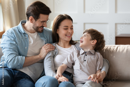 Happy young parents with little son cuddling, sitting on cozy couch, laughing father wearing glasses and mother having fun with preschool child boy, enjoying leisure time on weekend at home