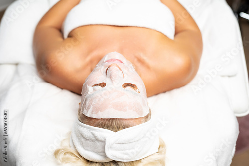 Blonde woman in a salon with a fabric moisturizing mask on her face. Caring for healthy skin.