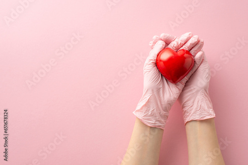 First person top-view perspective reveals female hands adorned in medical gloves, holding red heart that signifies cardiology. The pastel pink backdrop is perfect for your accompanying text or advert