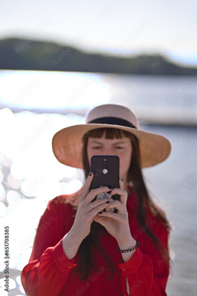 Kobieta w kapeluszu i czerwonej koszuli robiąca zdjęcie telefonem dla fotografa na tle jeziora w słoneczny letni dzień - obrazy, fototapety, plakaty 