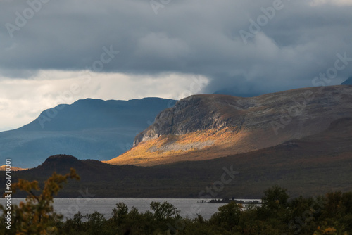 Sunny mountain side. Cloudy weather otherwise creating moody atmosphere. Lakeshore on the foreground. Norway region.