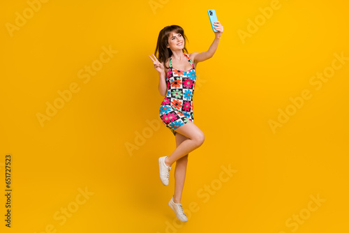 Full length photo of pretty cute lady wear flower print clothes jumping high recording video vlog v-sign isolated yellow color background