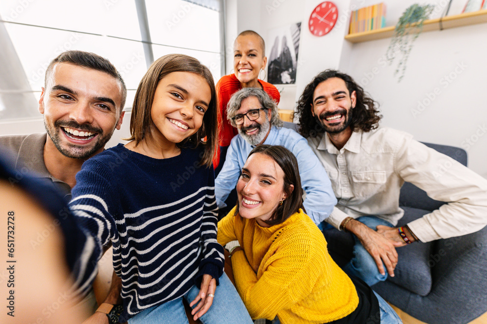 © Xavier Lorenzo - Happy self portrait of multi-generational family at home. Little cute girl taking selfie with parents, uncle, aunt and grandfather sitting together on sofa. Mobile phone screen on video call.