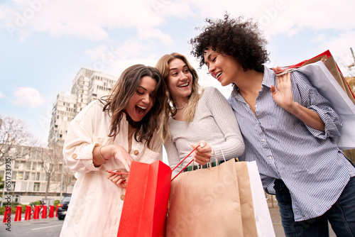 Three enthusiastic European friends on city street hold paper sack. Women coming from shopping in the sales look at inside the recyclable bags surprised. Smiling amazed people addicted to purchases. 