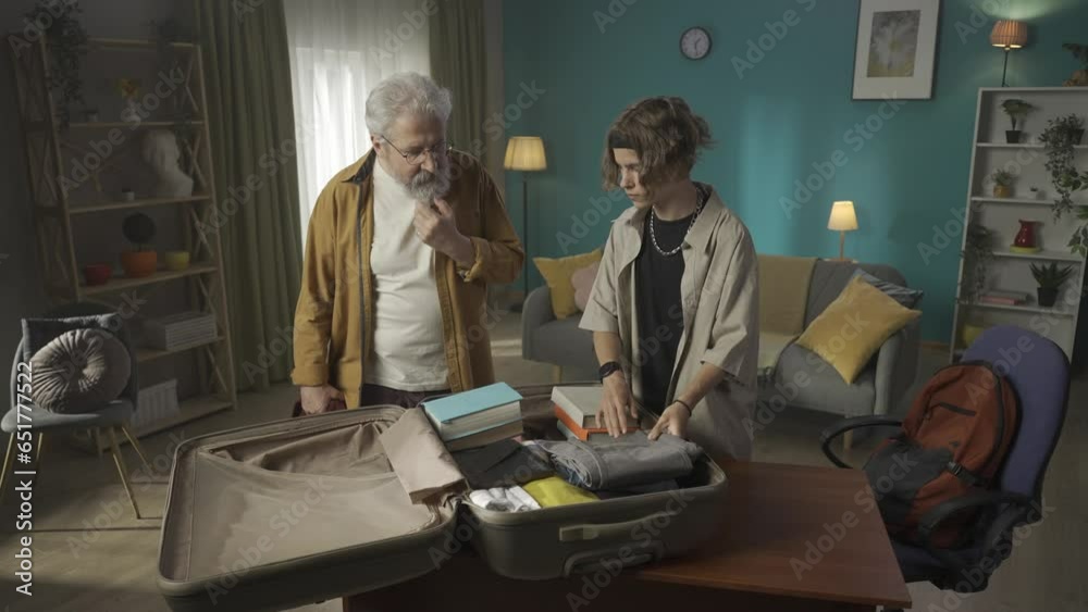 Video of a teenager, young man packing his belongings in a suitcase. His grandfather is insisting on him taking a sweater, but grandson refuses. HDR BT2020 HLG Material.