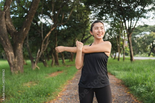 An Asian woman warms up her muscles before going jogging in a park full of trees.