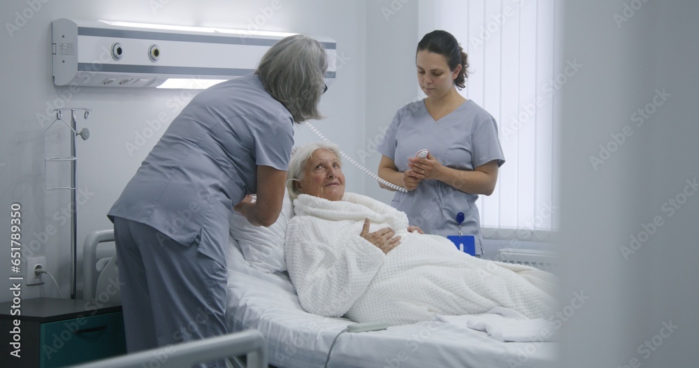 Mature nurse sets up bed using remote control, prepares hospital room for patient. Female medical worker pushes wheelchair with elderly woman and puts her in bed. Modern clinic or medical facility.