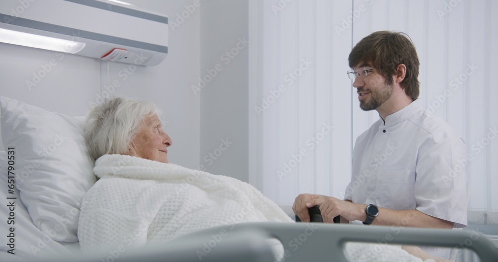 Doctor consults female elderly patient lying and resting in bed in hospital room. Male physician uses digital tablet, talks with nurse. Senior woman recovering after successful surgery. Modern clinic. Doctor consults female elderly patient lying and resting in bed in hospital room. Male physician uses digital tablet, talks with nurse. Senior woman recovering after successful surgery. Modern clinic.