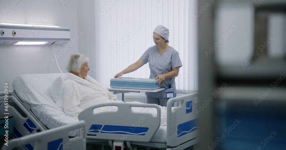 Nurse opens big lunch box with meal to female old patient. Elderly woman resting in bed in bright hospital room, recovering after successful surgery, eating food. Modern medical facility or clinic. Nurse opens big lunch box with meal to female old patient. Elderly woman resting in bed in bright hospital room, recovering after successful surgery, eating food. Modern medical facility or clinic.