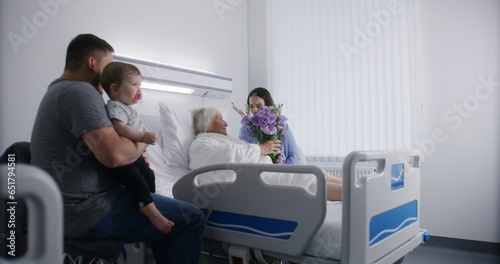 Elderly woman lying and resting in bed in bright hospital ward, plays with grandson. Loving family members support grandmother recovering after successful surgery. Modern medical facility or clinic.