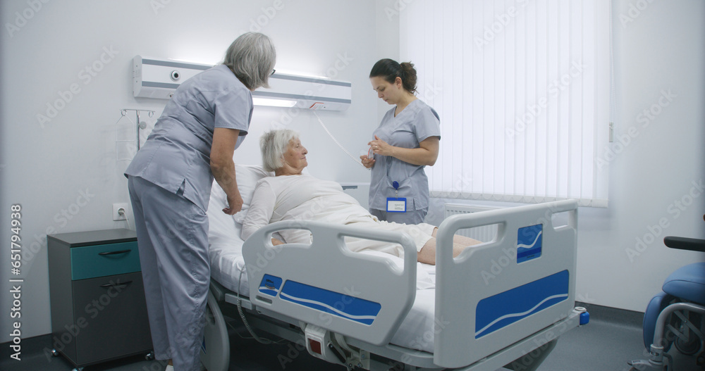 Mature nurse sets up bed using remote control, prepares hospital room for patient. Female medical worker pushes wheelchair with elderly woman and puts her in bed. Modern clinic or medical facility.