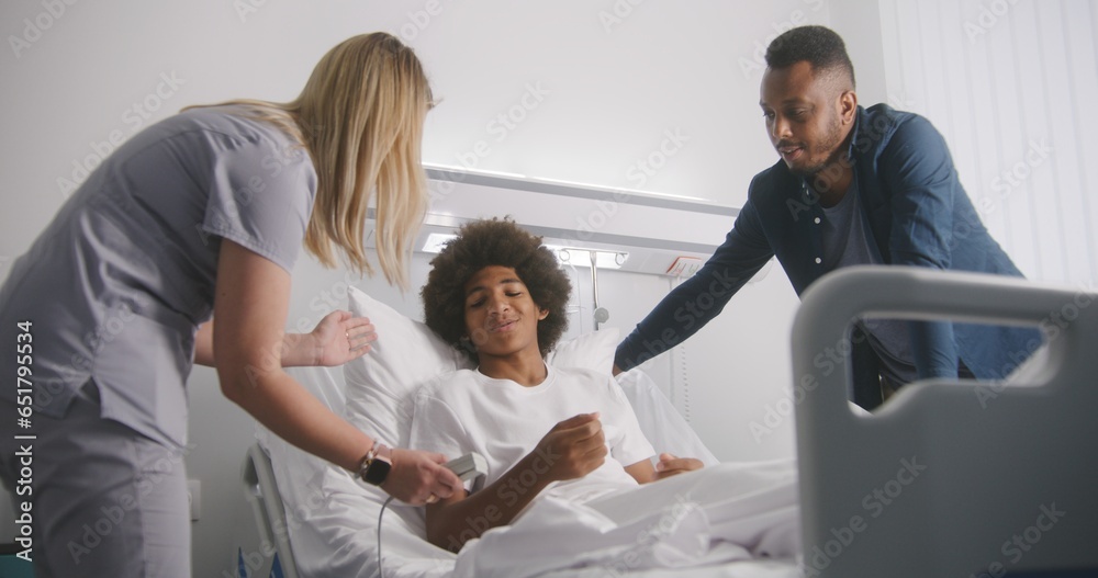 Man visits son in medical center and discusses results of operation. African American boy rests on bed in hospital ward. Patient fully recovering after successful surgery. Modern and bright clinic. Man visits son in medical center and discusses results of operation. African American boy rests on bed in hospital ward. Patient fully recovering after successful surgery. Modern and bright clinic.