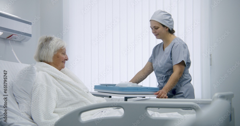 Nurse opens big lunch box with meal to female old patient. Elderly woman resting in bed in bright hospital room, recovering after successful surgery, eating food. Modern medical facility or clinic.
