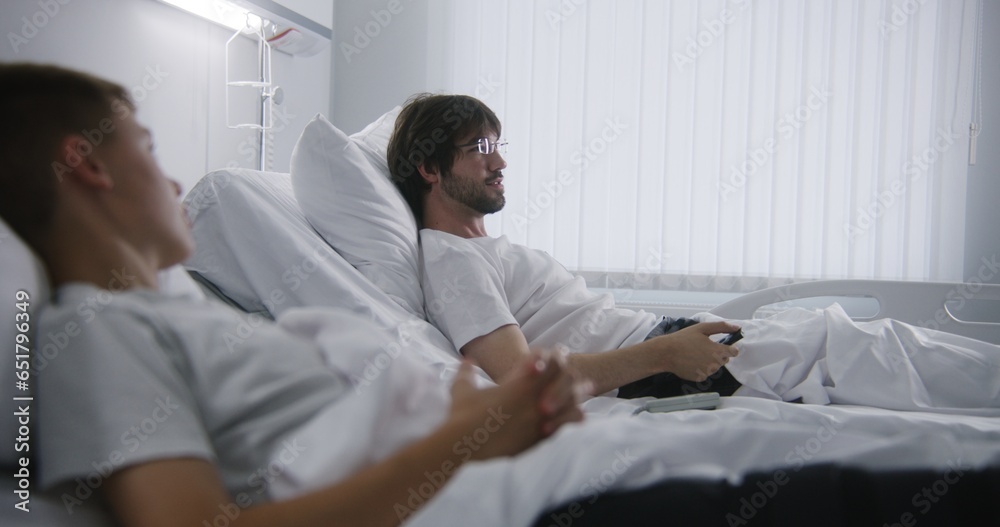 Obraz premium Adult man talks with kid, puts on glasses and turns on TV. Patients rest together in hospital ward. Hospital roommates fully recovering after successful operations. Modern and bright medical center.