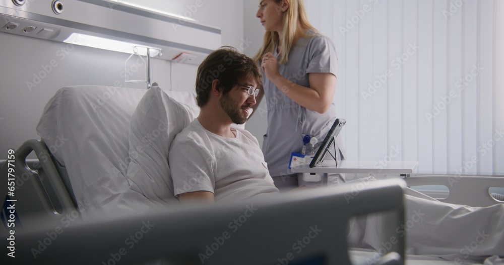 Adult man lies on bed in hospital ward, talks with partners on online conference using tablet. Man in glasses remotely works. Patient fully recovering after successful surgery. Modern bright clinic. Adult man lies on bed in hospital ward, talks with partners on online conference using tablet. Man in glasses remotely works. Patient fully recovering after successful surgery. Modern bright clinic.