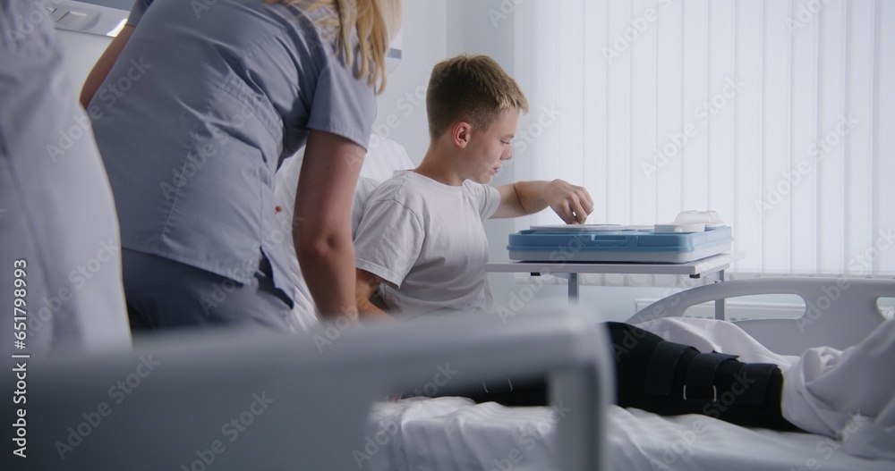 Nurse gives lunch box to resting patient and raises back of bed. Young boy fully recovering after successful operation in medical center. Modern bright clinic. Medical personnel work in hospital ward. Nurse gives lunch box to resting patient and raises back of bed. Young boy fully recovering after successful operation in medical center. Modern bright clinic. Medical personnel work in hospital ward.