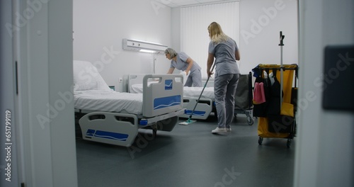 Adult cleaner mops floor with broom in hospital room. Mature health worker changes bedclothes after patients. Nurses clean hospital ward and talk. Medical staff at work in modern medical center.