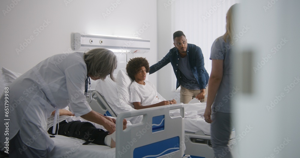 Obraz premium Mature physician bandages leg of young boy. African American man visits son in hospital ward. Nurse comes to teenager and raises back of his bed. Patient fully recovering after successful operation.