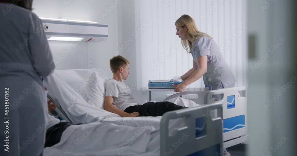 Nurse gives lunch box to resting patient and raises back of bed. Young boy fully recovering after successful operation in medical center. Modern bright clinic. Medical personnel work in hospital ward. Nurse gives lunch box to resting patient and raises back of bed. Young boy fully recovering after successful operation in medical center. Modern bright clinic. Medical personnel work in hospital ward.