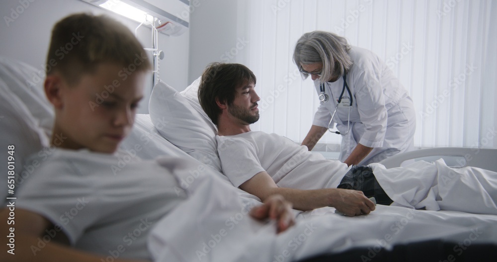 Obraz premium Man rests in bed, talks with young kid and turns on TV. Mature doctor does checkup of patients in hospital ward. Hospital roommates fully recovering after successful operations. Modern bright clinic.
