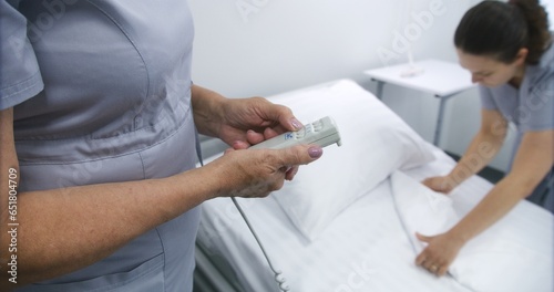 Mature nurse adjusts smart bed in hospital ward using lifting remote control. Another female medical worker prepares bedding for patient. Modern equipment in clinic or medical facility. Close up.