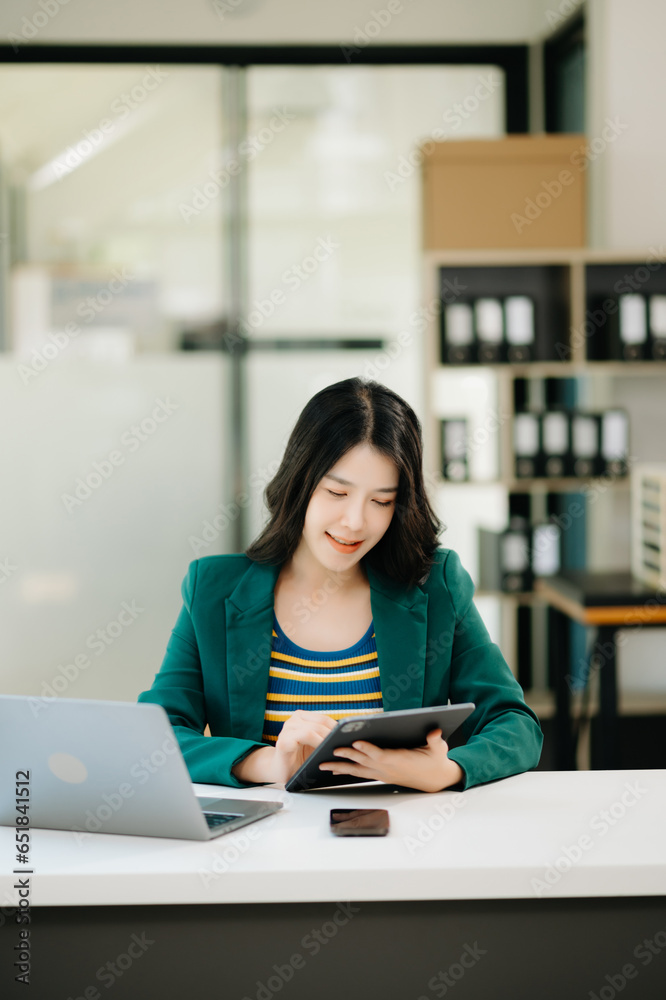 Fototapeta premium Beautiful Asian woman using laptop and tablet while sitting at her working place. Concentrated at work. in office..