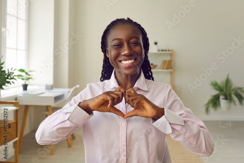 Happy business woman shows her love and gratitude. Video call headshot view thankful African American lady in shirt sitting in home office, looking at camera, smiling, making heart shape with hands photo