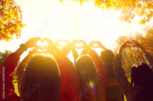 Group of people greeting sunrise in nature. Young adult male and female friends standing in autumn park at dawn, holding up hands and forming heart shapes in bright sunlight. Back view from behind