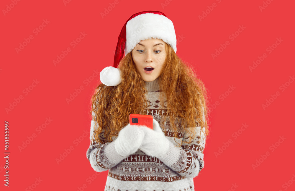 Cute girl in winter clothes is amazed saying wow omg while reading message with good news. Young woman in Santa hat and Christmas sweater looking at smartphone screen in amazement on red background.