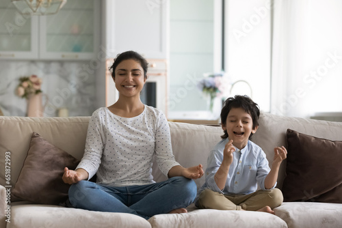 Smiling Indian mother with 5s son meditating on couch together, sitting in easy seat lotus, happy young mom teaching little boy child to practicing yoga, healthy lifestyle, enjoying leisure time
