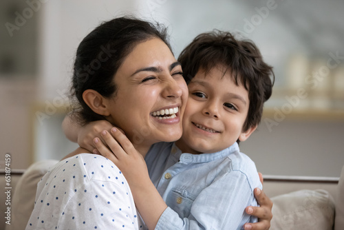Head shot close up of excited funny Indian mother hugging adorable 5s son, happy laughing young mom and child boy enjoying tender moment, cuddling, good relationship, sitting on couch at home